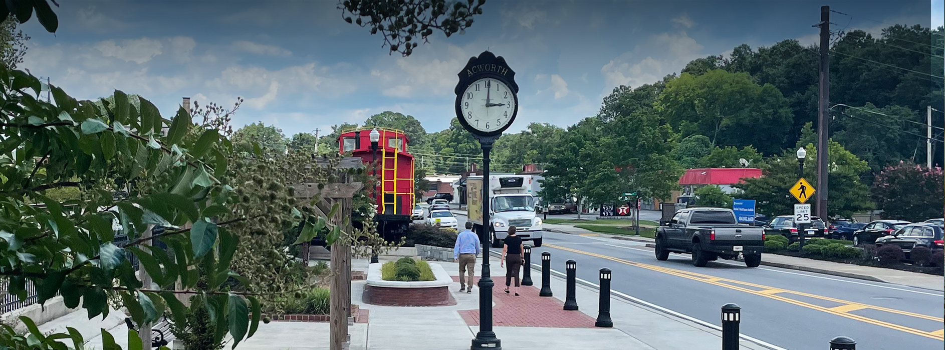 Sidewalk scene with people walking near a clock post and a railroad car, adjacent to a busy street with passing vehicles and lush green trees in the background.