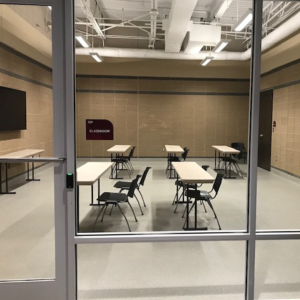 A classroom with beige walls and a glass door, equipped with six small tables and chairs placed in pairs, and a wall-mounted TV screen on the left. The room is empty.