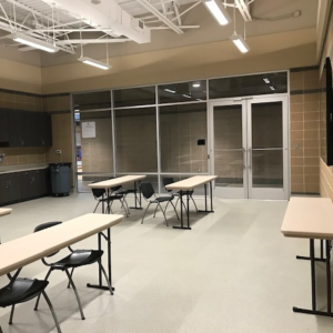 A classroom with beige walls and a tiled floor features two rectangular tables and four black chairs. Glass doors and windows are at the back of the room, allowing visibility into an adjacent area.