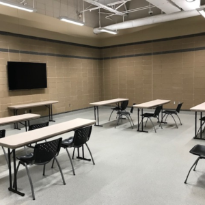 A classroom setting with multiple tables and chairs arranged in a spaced-out manner, a wall-mounted TV, and beige walls.