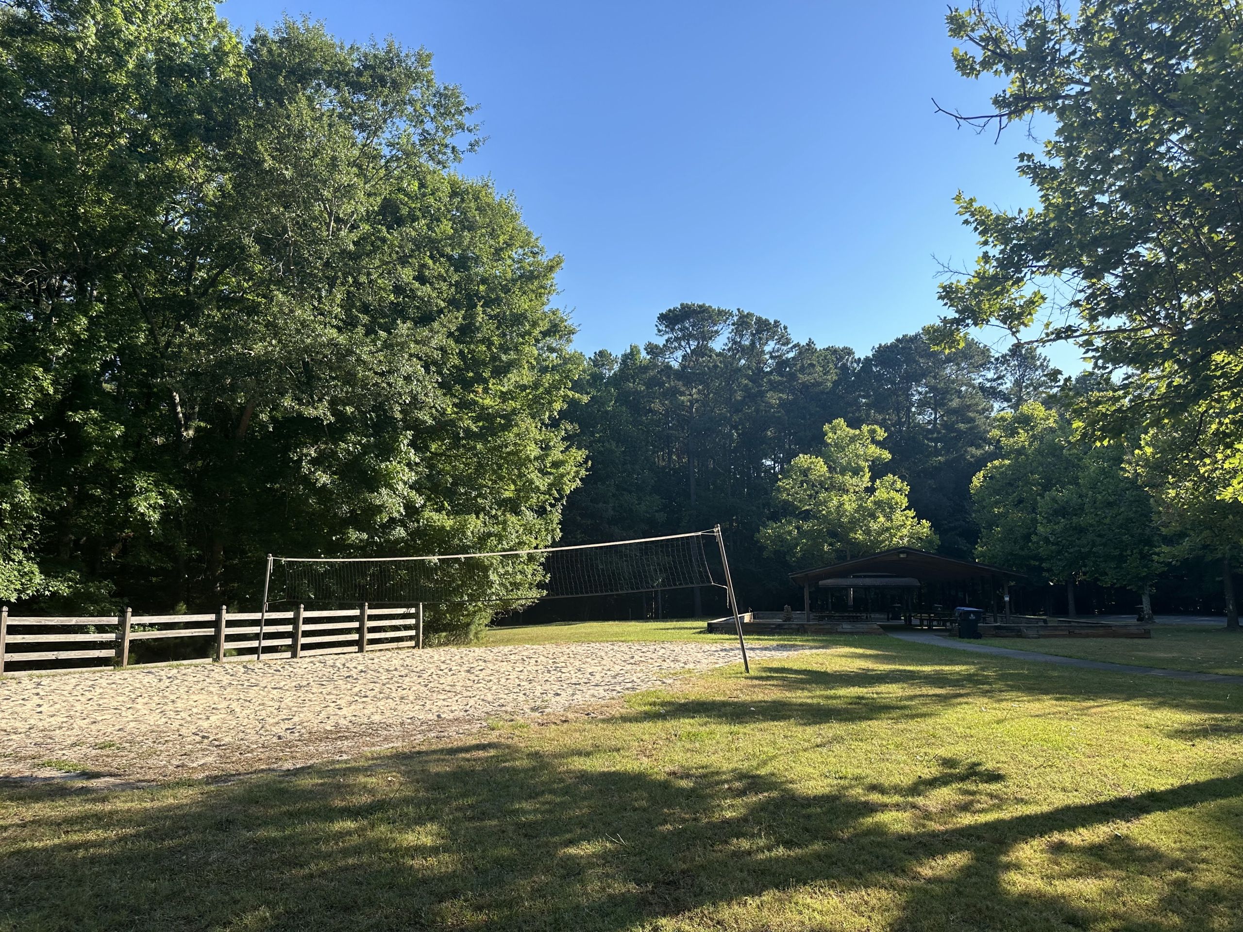 image A volleyball court surrounded by trees and a fenced area with a pavilion in the background under a clear blue sky.