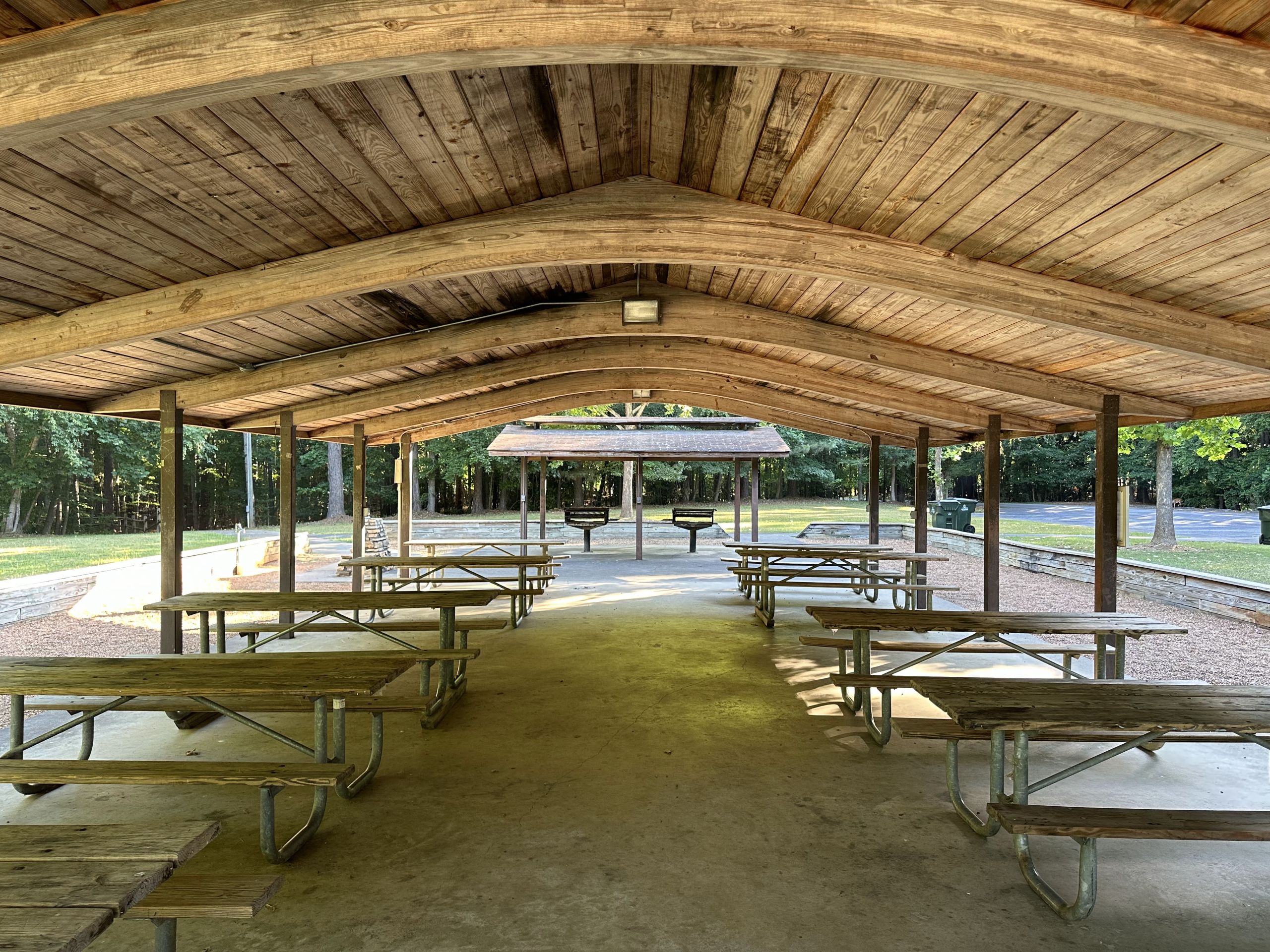 image A spacious, open-air pavilion with wooden roof and multiple picnic tables is situated in a park setting, with green trees in the background.