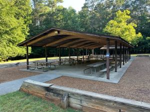 image A wooden pavilion with picnic tables underneath, located in a park with surrounding trees and open grassy areas.