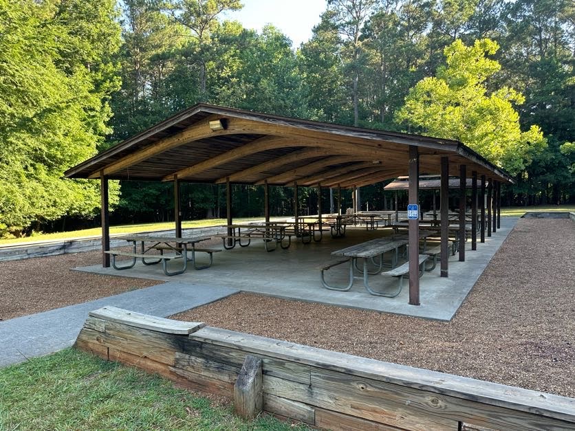 image A wooden pavilion with picnic tables underneath, located in a park with surrounding trees and open grassy areas.