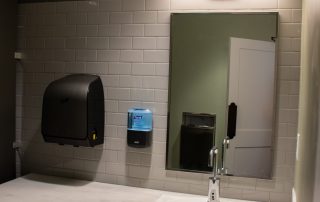 A public restroom sink area with a white basin, soap dispenser, and hand dryer beneath an illuminated mirror against a tiled wall.
