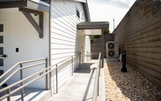 Accessible pathway with handrails next to a building and air conditioning units, supported by a retaining wall with pebbles. Steps and a lamppost are visible in the background.
