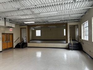 An empty, well-lit hall with a small stage at the back, accessible via steps on both sides. There are doors to the left and large windows on the right. The ceiling has exposed beams and lights.