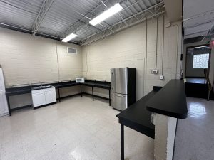 A minimalist kitchen with a sink, microwave, stainless steel refrigerator, and long black countertops. The room has beige walls, a white tiled floor, and industrial-style ceiling with exposed pipes.