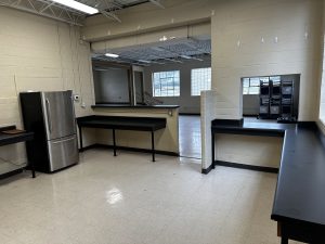 A room with beige walls, industrial lighting, and black countertops; features a stainless steel refrigerator and several empty shelves. An open doorway leads to another room with large windows.