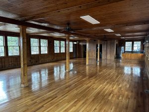 An empty room with polished wooden floors, wooden walls, exposed wooden beams, and large windows on one side. Several light fixtures hang from the ceiling.