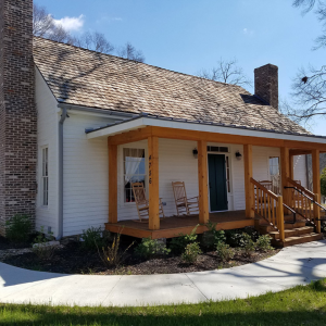 A small white house with a brick chimney and a wooden porch featuring two rocking chairs. The house number "4756" is visible on the porch post.