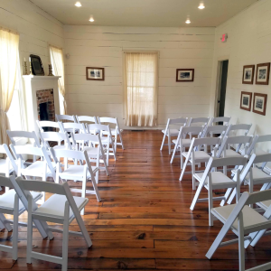 A small room with wooden flooring, white wooden walls, and several rows of white folding chairs facing a central area. Framed pictures hang on the walls, and there is a brick fireplace on the left side.