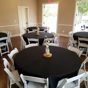 A small event room with round tables covered in black tablecloths and white chairs. Each table has a simple floral centerpiece. The room has light-colored walls, wood floors, and an open door to the outside.