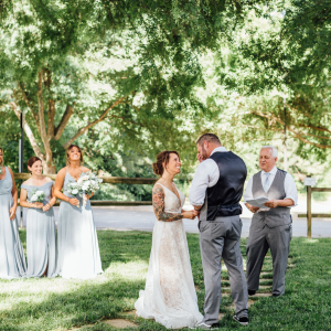 A couple exchanges vows during an outdoor wedding ceremony. Bridesmaids in gray dresses stand to the left, while an officiant stands to the right under a canopy of trees.