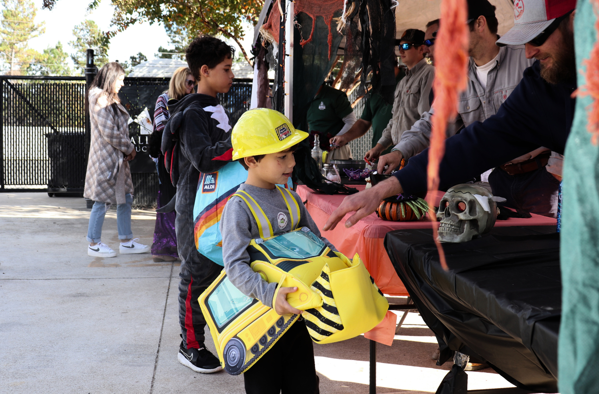 A child in a construction costume stands at an outdoor booth, receiving candy from an adult during a Halloween event; other people and decorations are visible in the background.