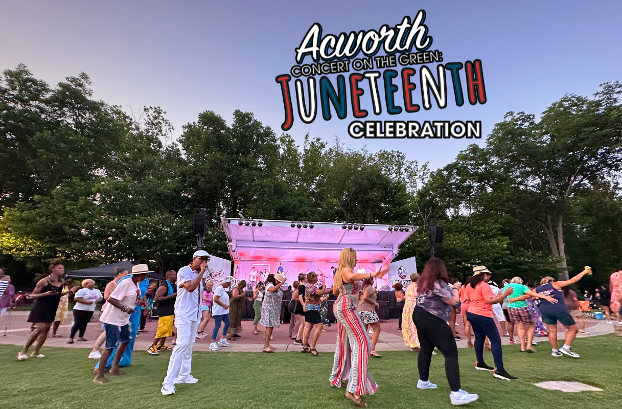 People dance on a grassy lawn in front of a stage at the Acworth Concert on the Green Juneteenth Celebration, with trees and vibrant event signage overhead, capturing the joyful spirit of Juneteenth.