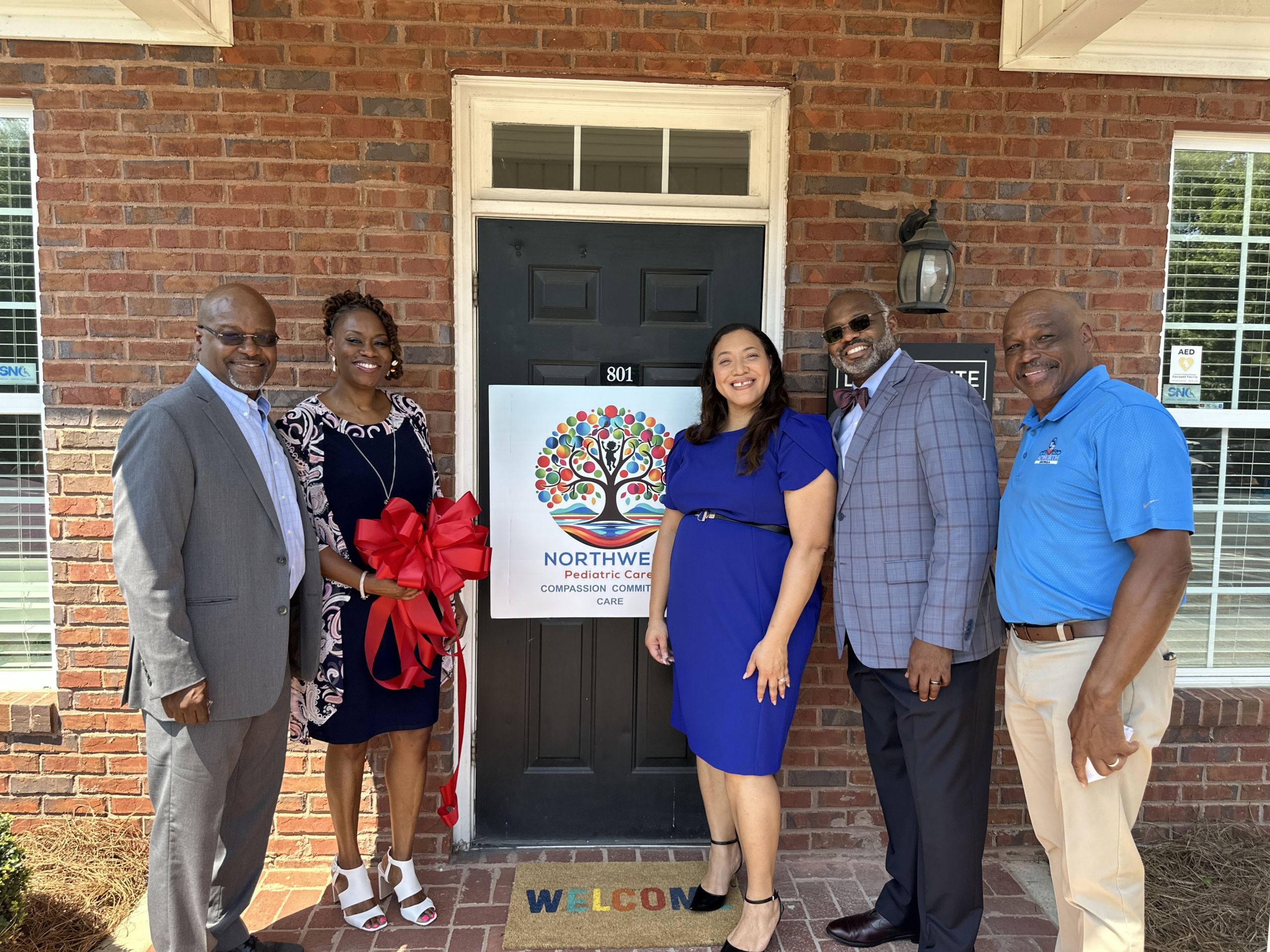 Five people stand in front of a brick building with a sign that reads "Northwest Metro Pediatric Care". They smile and pose around a door, with one person holding a large red ribbon.