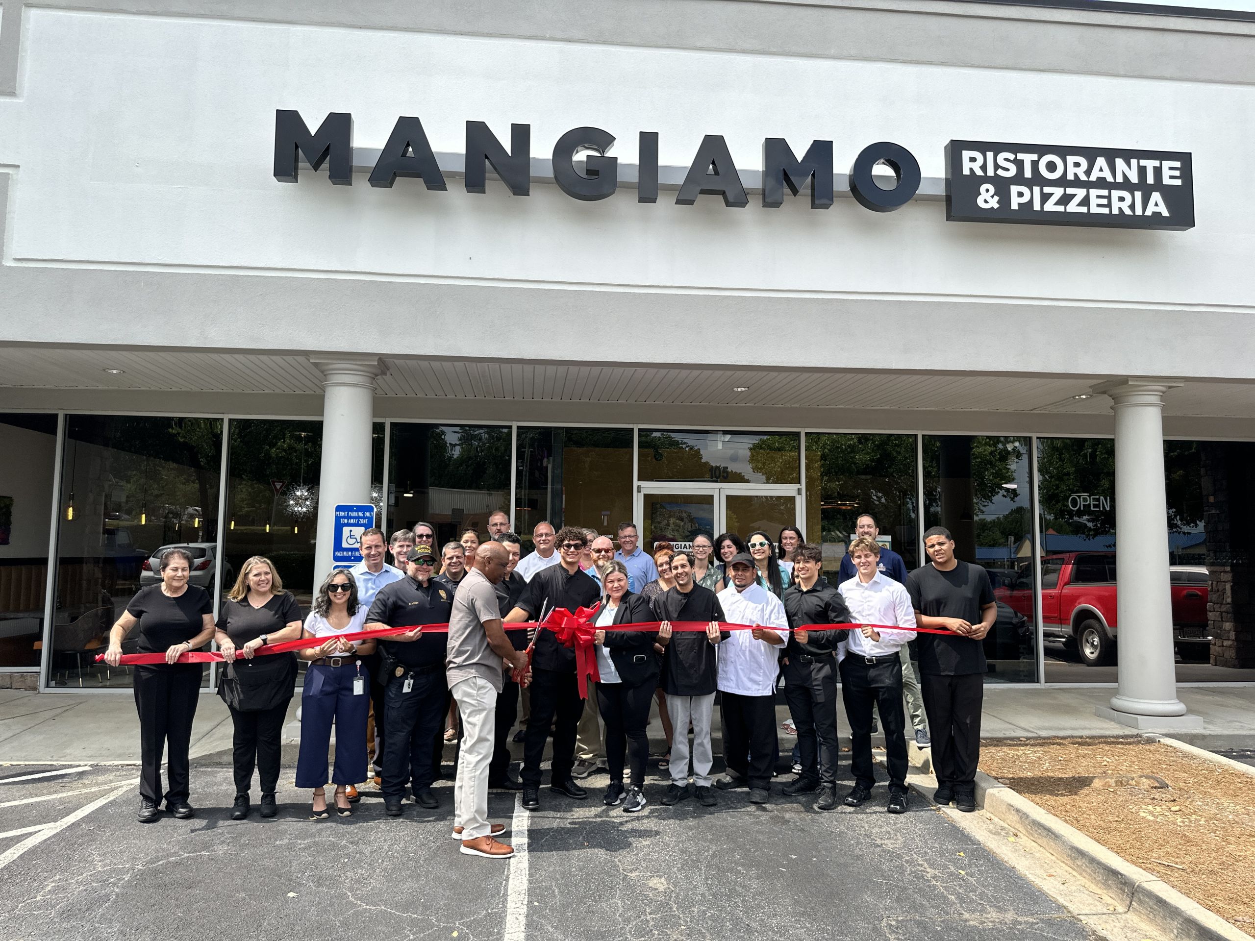 A group of people stands in front of "Mangiamo Ristorante & Pizzeria," holding a large red ribbon, suggesting a ribbon-cutting ceremony outside the restaurant.