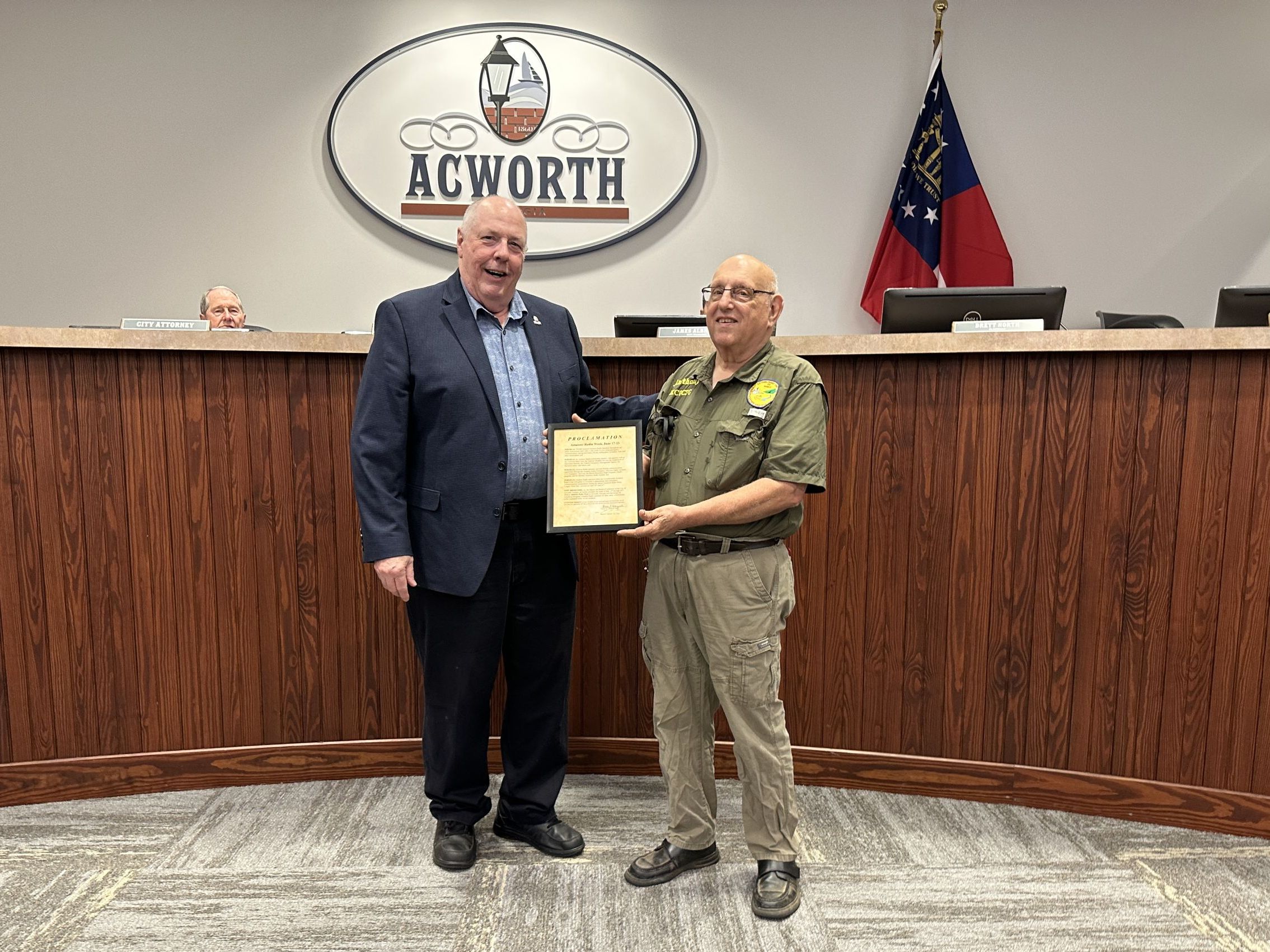 Two men stand in front of a wooden desk with the Acworth insignia. One man, in a suit, presents a plaque to the other man, who is wearing a green uniform. Additional people are seated behind the desk.