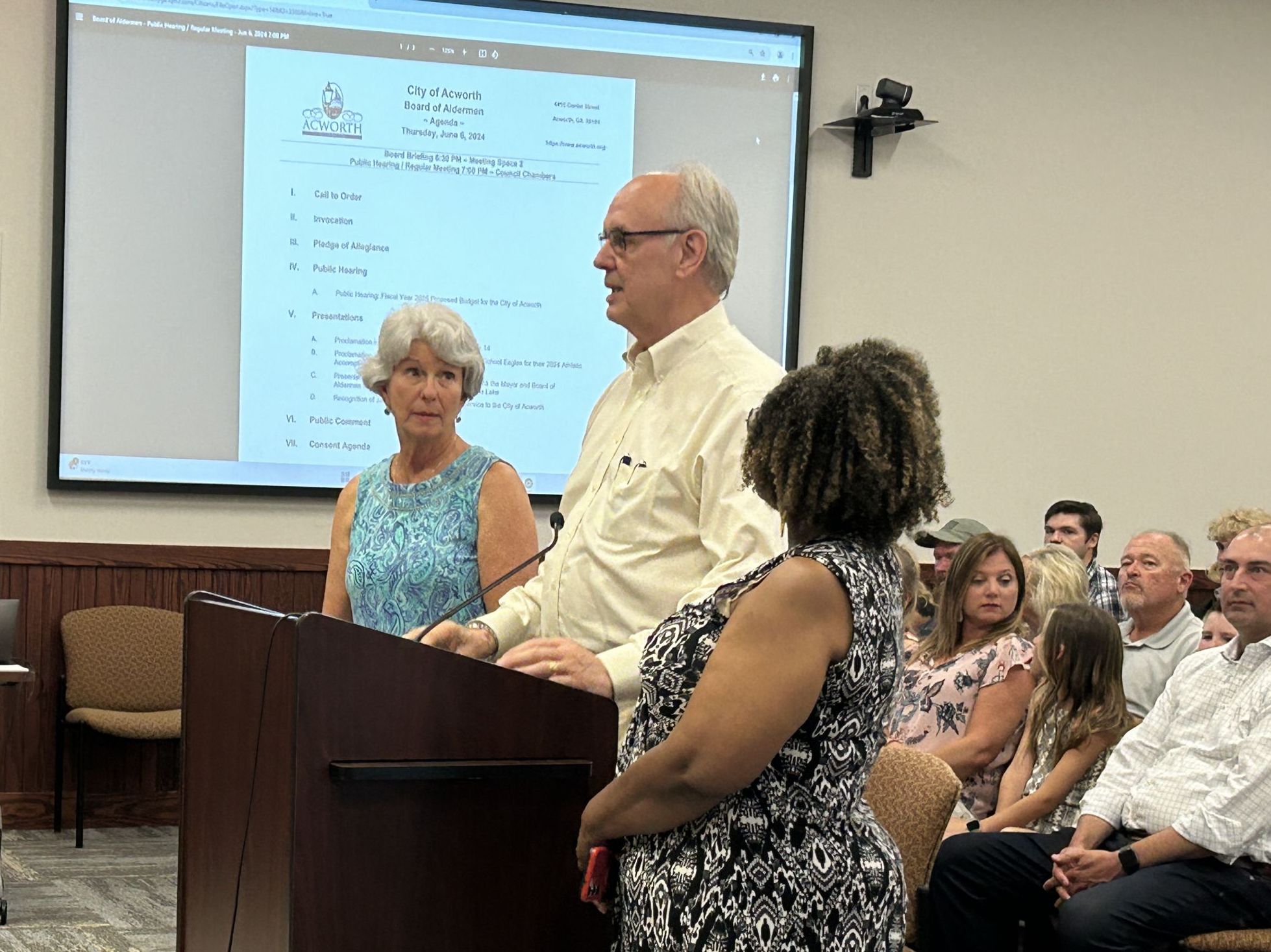 Three people stand at a podium in a meeting room, addressing an audience. The projector screen behind them displays an agenda for a city council meeting. Seated individuals watch attentively.