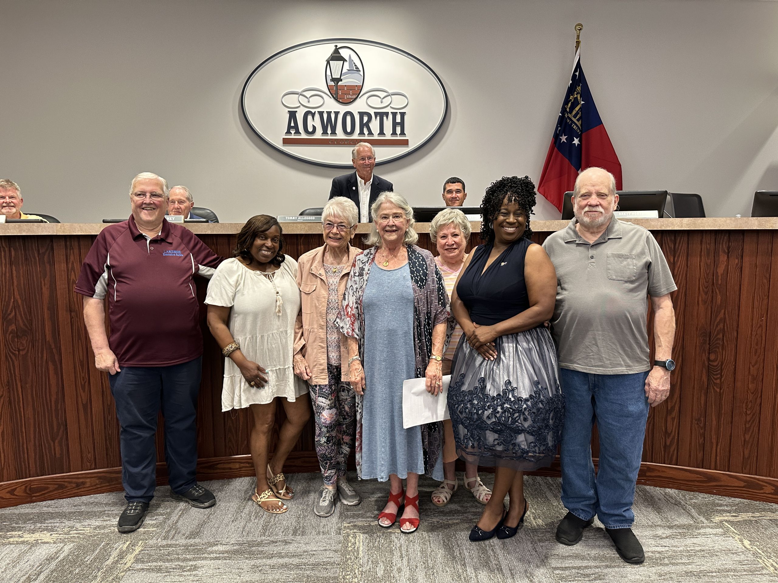 A group of people stands together in front of an "Acworth" sign, with a few individuals seated behind them at a wooden desk. An American flag is visible in the background.