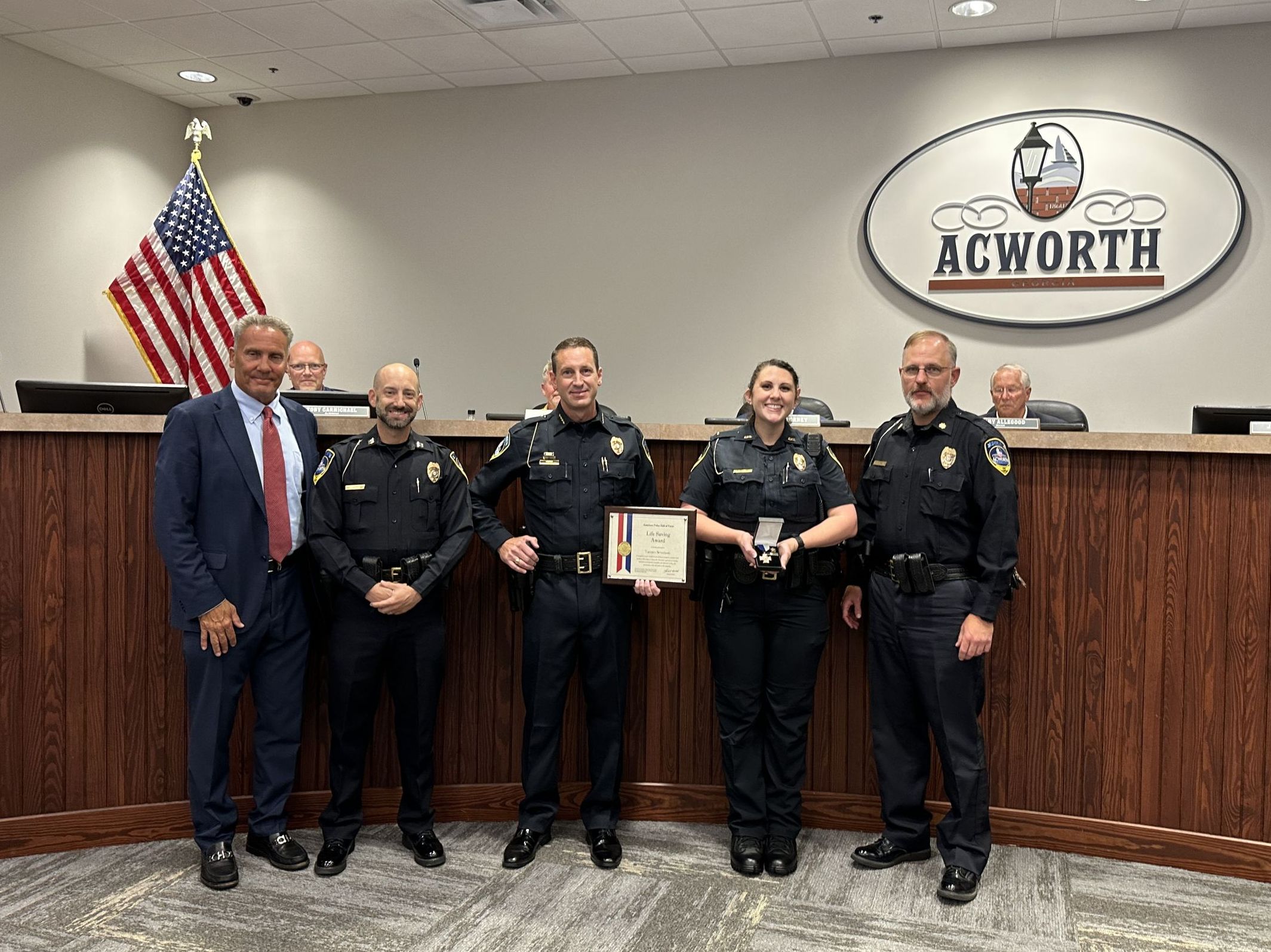 A group of individuals, including police officers, stands in front of a wooden podium with the Acworth logo. Two officers hold an award, while an American flag is displayed in the background.