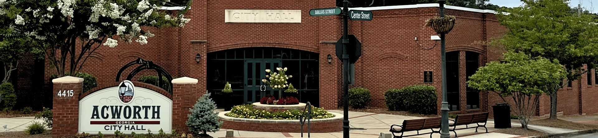 Front view of Acworth City Hall in Georgia, with a brick facade, entrance sign, and surrounding greenery. Street signs for Main Street and Cherokee Street are visible in the foreground.