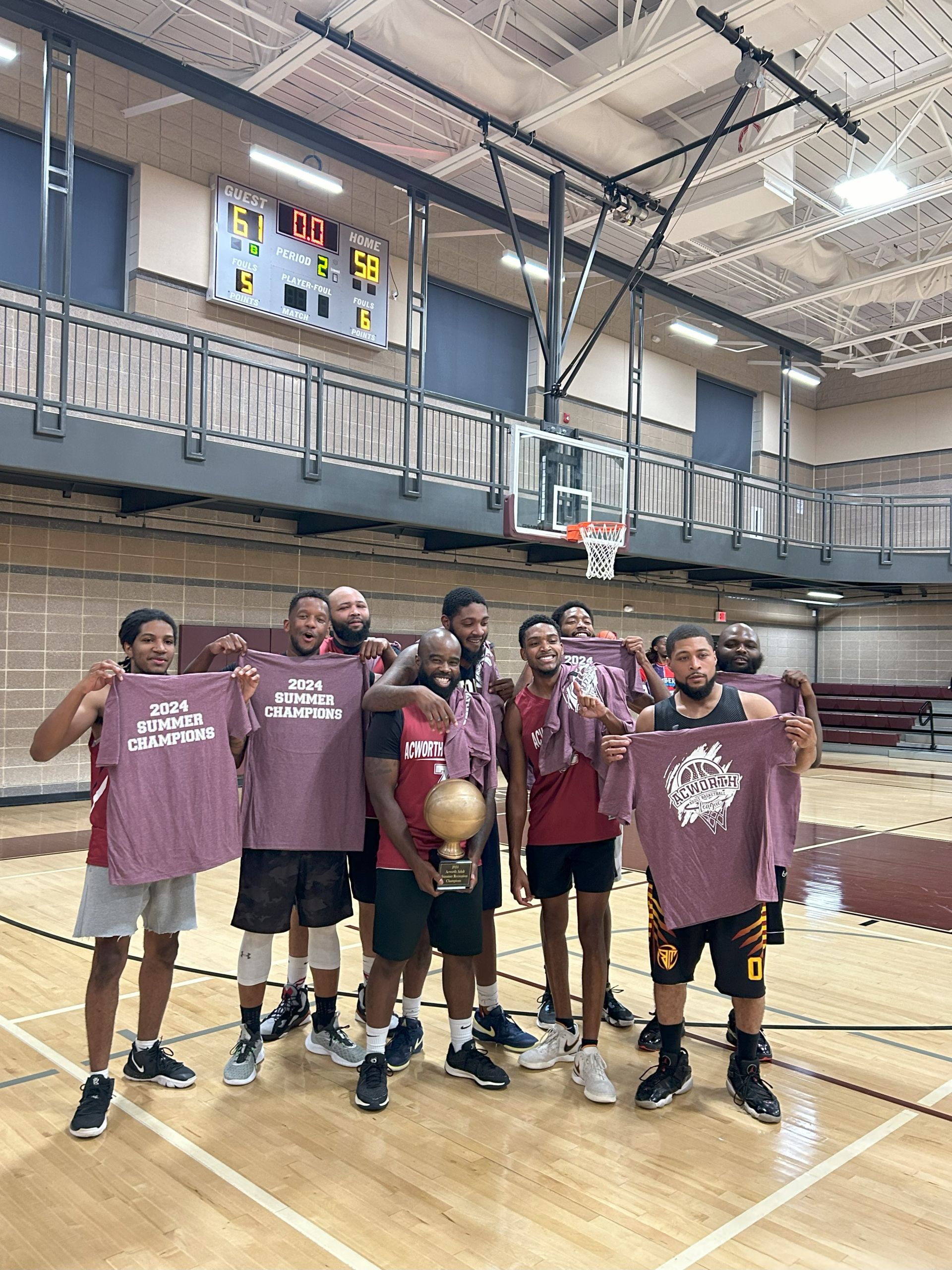 A basketball team stands together on an indoor court, holding "2024 Summer Champions" shirts and a trophy. The scoreboard behind them shows a final score of 61-58.