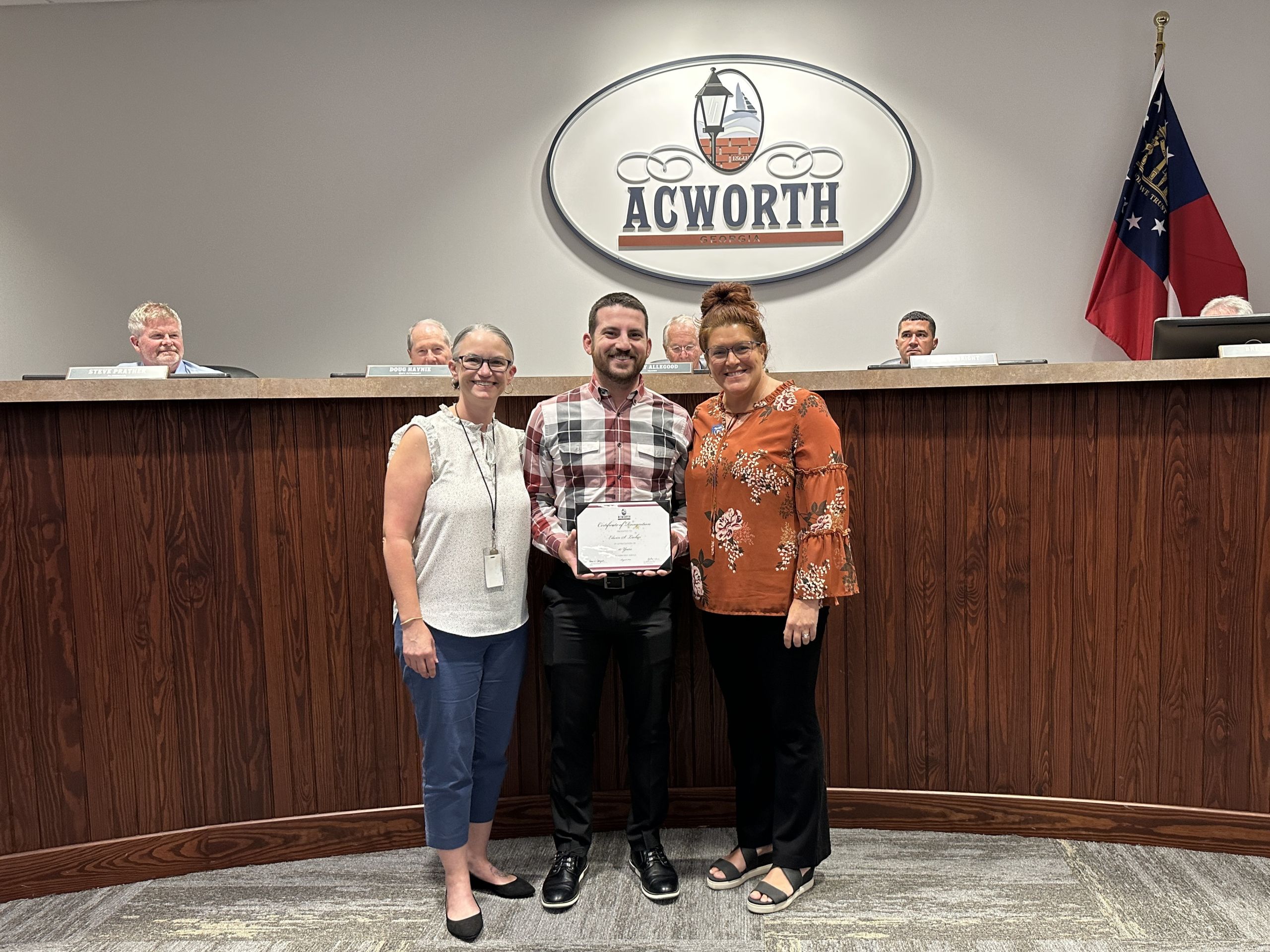 Image Three People posing for a photograph at Acworth City Council Chambers. Two women stand on outside and man in the middle is holding a certificate.