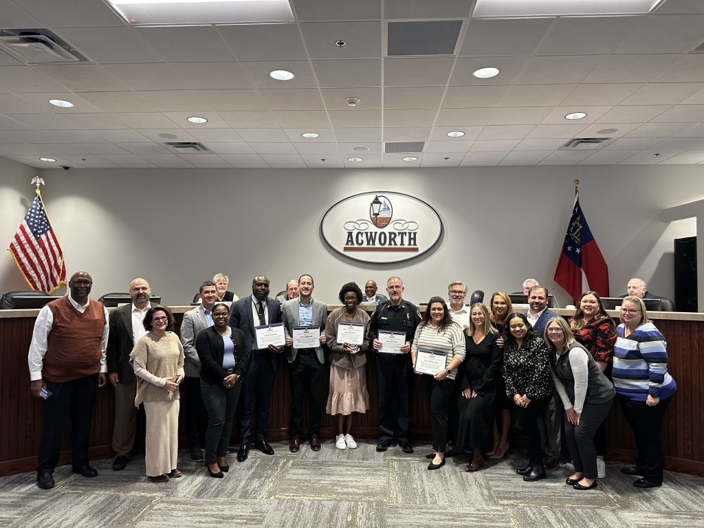 A group photo of individuals in an office setting, with some holding certificates. An "Acworth" sign is in the background. An American flag and another flag are visible.