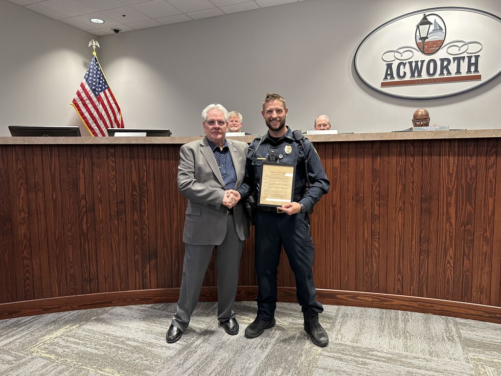 A man in a suit shakes hands with a uniformed officer holding a plaque. They stand in front of a wooden podium with an "ACWORTH" sign and an American flag. Four people are seated behind.