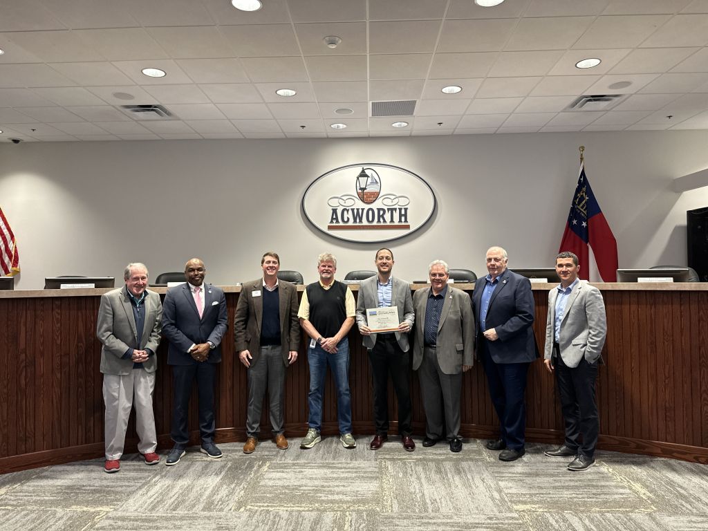 A group of eight people stands in front of a wooden podium and logo that reads "Acworth," with one person holding a certificate.