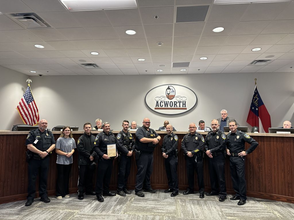 A group of law enforcement officers and officials stand together in a meeting room, with the Acworth logo on the wall behind them.