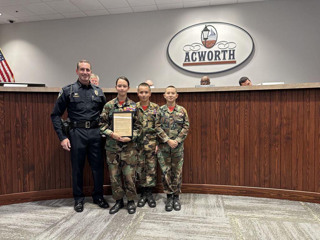 A police officer stands with three youth cadets in military uniforms holding a certificate, in a room with an "Acworth" sign.