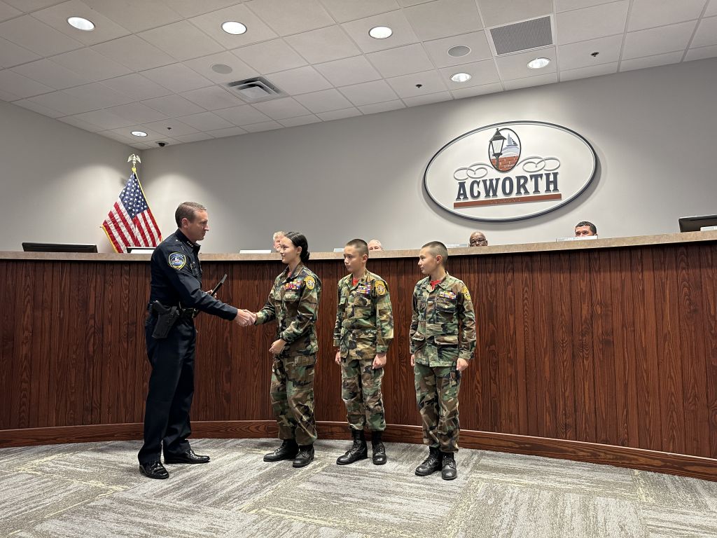 A police officer shakes hands with a woman in uniform, standing with two youths in similar uniforms at a podium, with an American flag and an "Acworth" sign in the background.