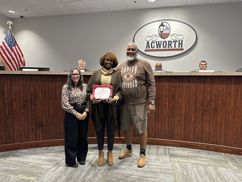 Three people stand in front of a wood-paneled desk; the middle person holds a framed certificate. A logo on the wall reads "Acworth," and a U.S. flag is visible to the left.