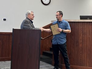 Two men shaking hands. One holds a framed certificate. A clock is on the wall above them.