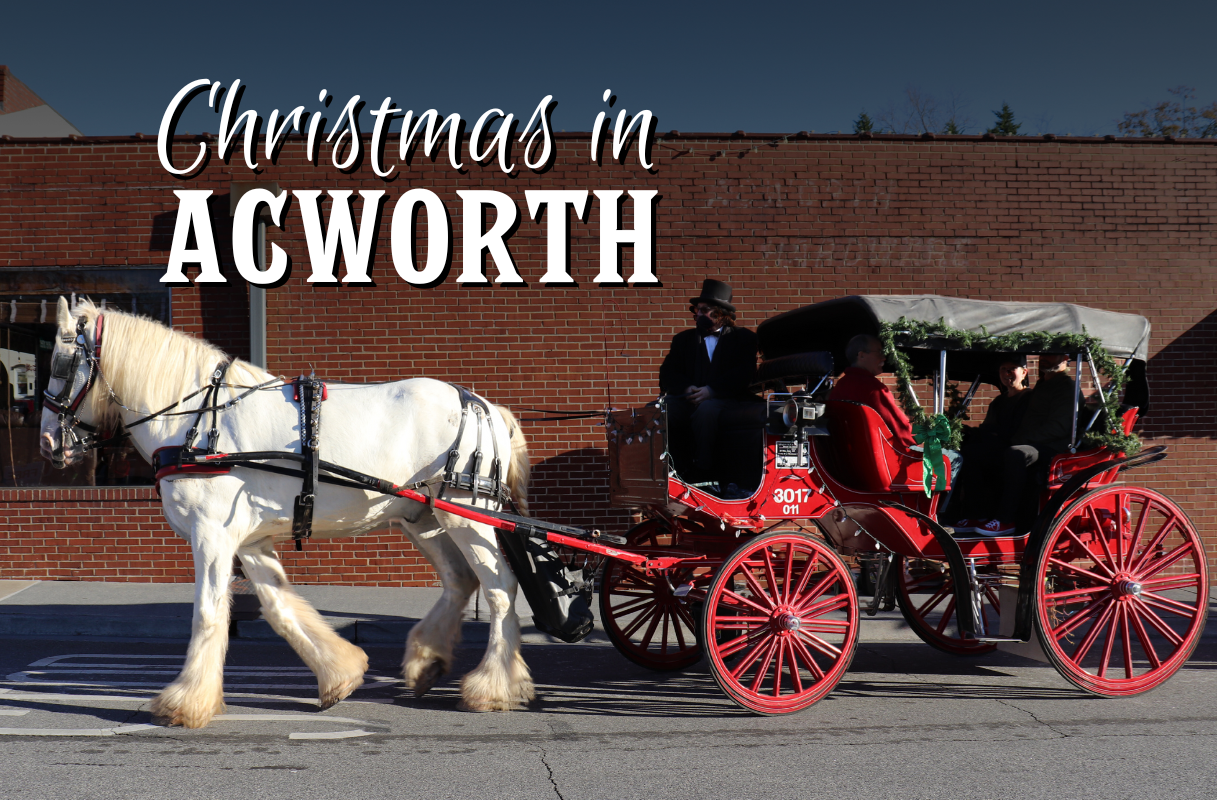 A horse-drawn carriage with people in festive attire passes by a brick building, with "Christmas in Acworth" text above.