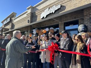 A group of people gather for a ribbon-cutting ceremony outside a Bagel Run store. Balloons decorate the entrance.