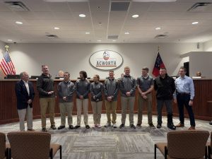 A group of nine people stands in a row at a ceremony in a conference room with a seal of "Acworth" on the wall. Two flags are visible in the background.