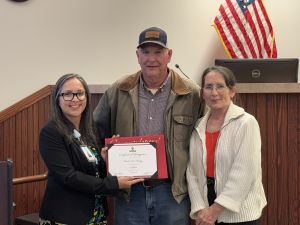 Three people pose with a certificate in a room. An American flag and a computer are visible in the background.