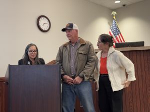 Three people stand behind a podium in a room with a clock and an American flag in the background.