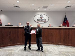 A police officer and a man stand holding a certificate in a council chamber with "Acworth" on the wall. Seven people are seated at a long desk in the background.