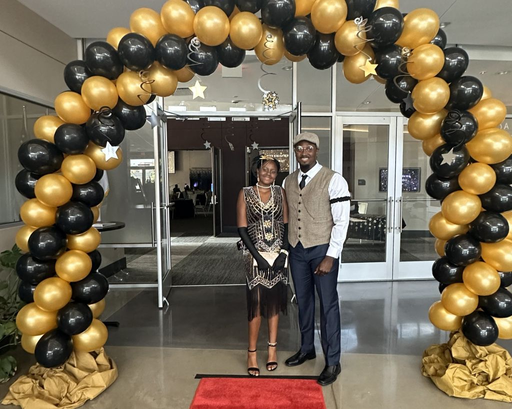 Two people dressed in formal attire stand under a black and gold balloon archway on a red carpet at a Daddy Daughter Date Night event in an indoor setting.