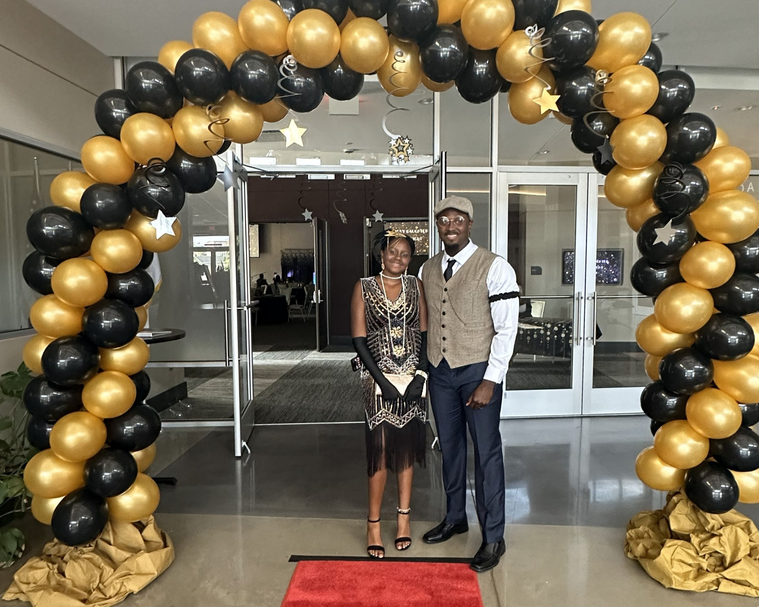 Two people dressed in formal attire stand under a black and gold balloon archway on a red carpet at a Daddy Daughter Date Night event in an indoor setting.