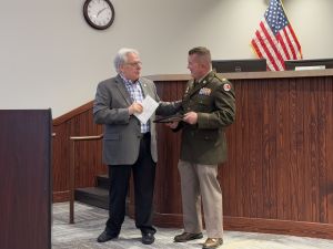 Two men stand in an office setting; one in a gray suit, the other in a military uniform. They appear to be exchanging documents. An American flag and a clock are in the background.