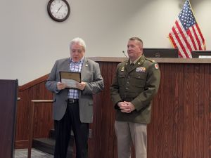 Two men stand in a room; one in a suit holding a plaque, the other in a military uniform. An American flag and clock are in the background.