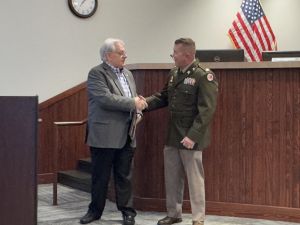 Two men shaking hands in a room with a U.S. flag and a clock on the wall. One man is in a suit, the other in a military uniform.