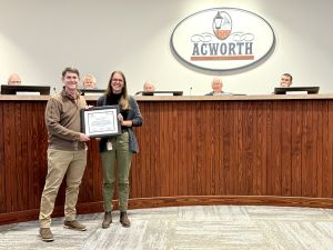 Two individuals stand holding a framed certificate in front of a council with an Acworth sign behind them.