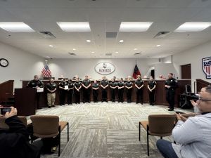 A group of uniformed officers stands in a line inside a room with a podium and seating area. A person on the right speaks, and a city emblem is visible on the wall behind them.